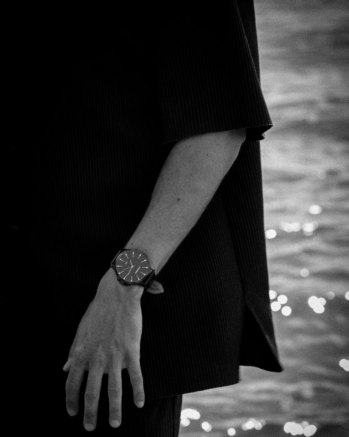 A wrist and watch against a dark coat with water behind, black and white