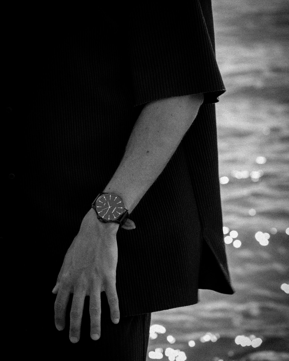 A wrist and watch against a dark coat with water glittering behind